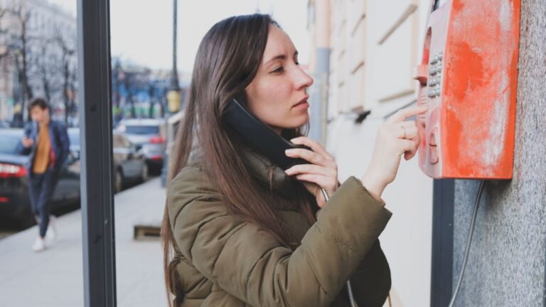 Young Woman Traveler Using Old Fashioned Public Telephone Metal Payphone Operated With Coins For Having Conversation