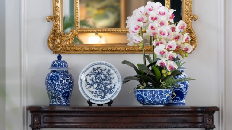 Blue paint porcelain collection with Orchid vase on a dark wooden table with golden mirror on white wall.