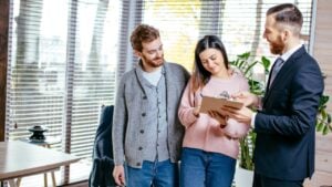 Confident male estate agent meeting with young couple in appartment for sale, discussing property investment with broker, construction company manager showing build project, apartment plan on papers.