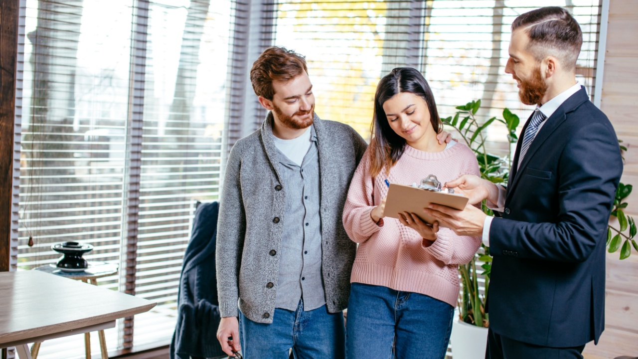 Confident male estate agent meeting with young couple in appartment for sale, discussing property investment with broker, construction company manager showing build project, apartment plan on papers.