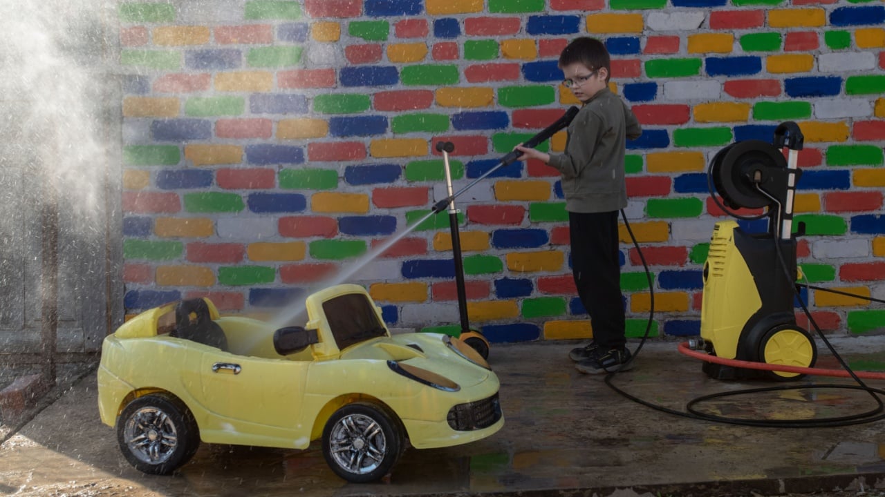 Happy child washes yellow toy car from high pressure washer in front of a multi-colored brick wall. Fun during the summer holidays. The concept of family business.