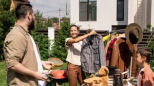 Positive young woman holding jacket on hanger and offering to buy it to husband at summer garage sale