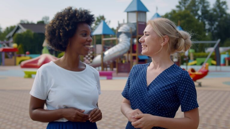 Medium shot of multiethnic young women talking and watching children play on playground. Portrait of african and caucasian mothers chat at park