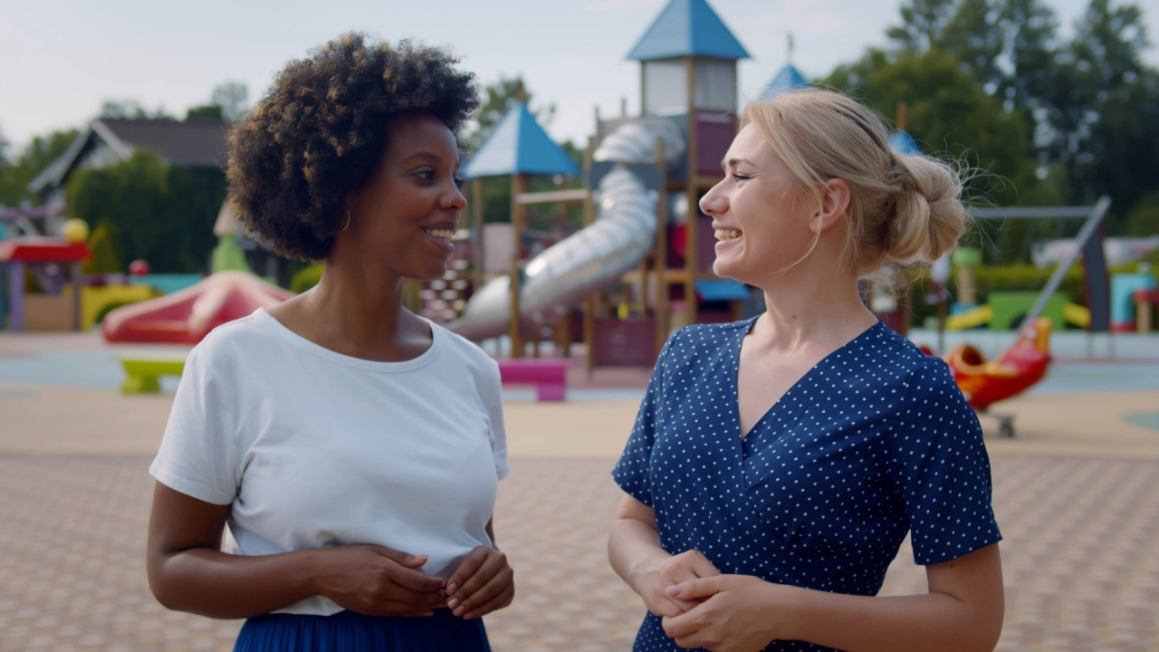 Medium shot of multiethnic young women talking and watching children play on playground. Portrait of african and caucasian mothers chat at park
