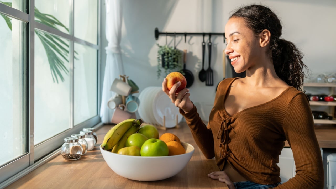 Young attractive Latino woman eat fruits on table in kitchen at home. Beautiful active female stand with happy face, enjoy eating apple, healthy foods for health care wellness to lose weight in house