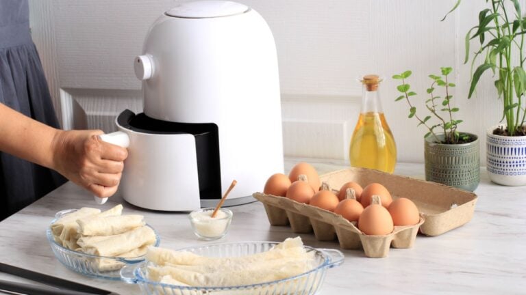 Woman Hand Open Airfryer Tray. A White Deep Fryer or Oil Free Fryer Appliance, Tongs, Clear Baking Dish and Egg Tray are on the Wooden Table in the Kitchen with a Small Plant in the Pot (air fryer)