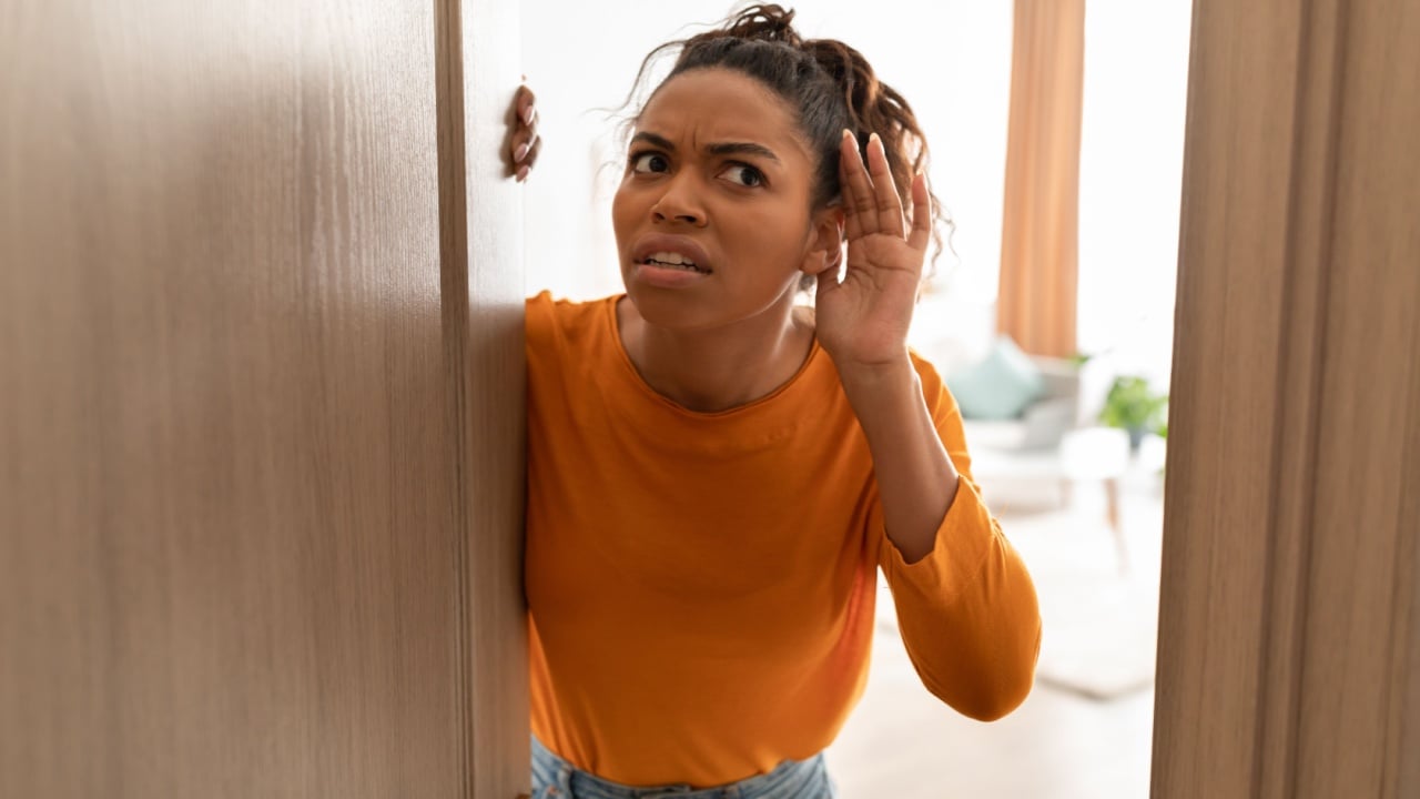 Displeased Black Lady Listening Holding Hand Near Ear Standing In Opened Door Having Problem With Soundproof At Home. Concerned Woman Hearing Something Openind Door Of Her House