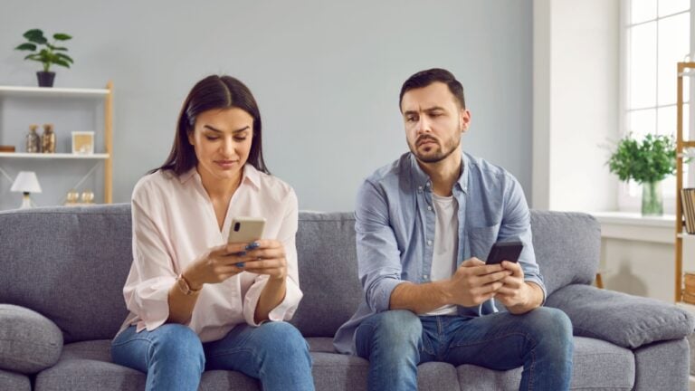 Jealous man spying looking at his girlfriend phone. Couple sitting on couch with smartphones, suspicious husband trying to read messages at wife smartphone while she texting message and smiling