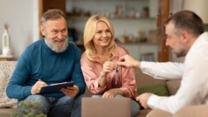 Real Estate Agent Giving New House Keys To Happy Mature Couple Buying Property And Signing Papers Sitting On Sofa Indoors. Apartment Rent And Purchase. Selective Focus On Spouses