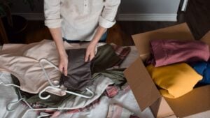 Woman is sorting through her clothes, folding and packing unwanted items into cardboard box for donations, top down view