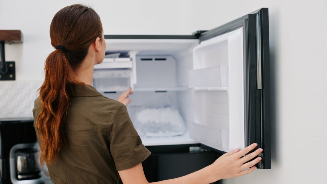 A woman in the kitchen of her home opened an empty freezer with ice inside, home refrigerator, defrosted, view from the back.