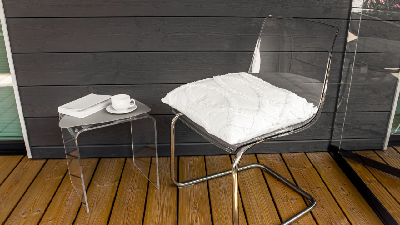 Clear transparent modern acrylic furniture in home. Chair with pillow, side table with tea cup and book.