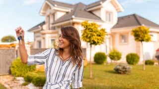 Beautiful young woman standing in front of her new house, holding the keys of her newly purchased property