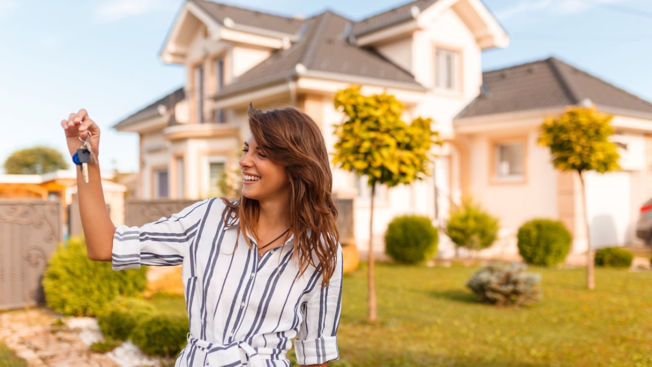 Beautiful young woman standing in front of her new house, holding the keys of her newly purchased property