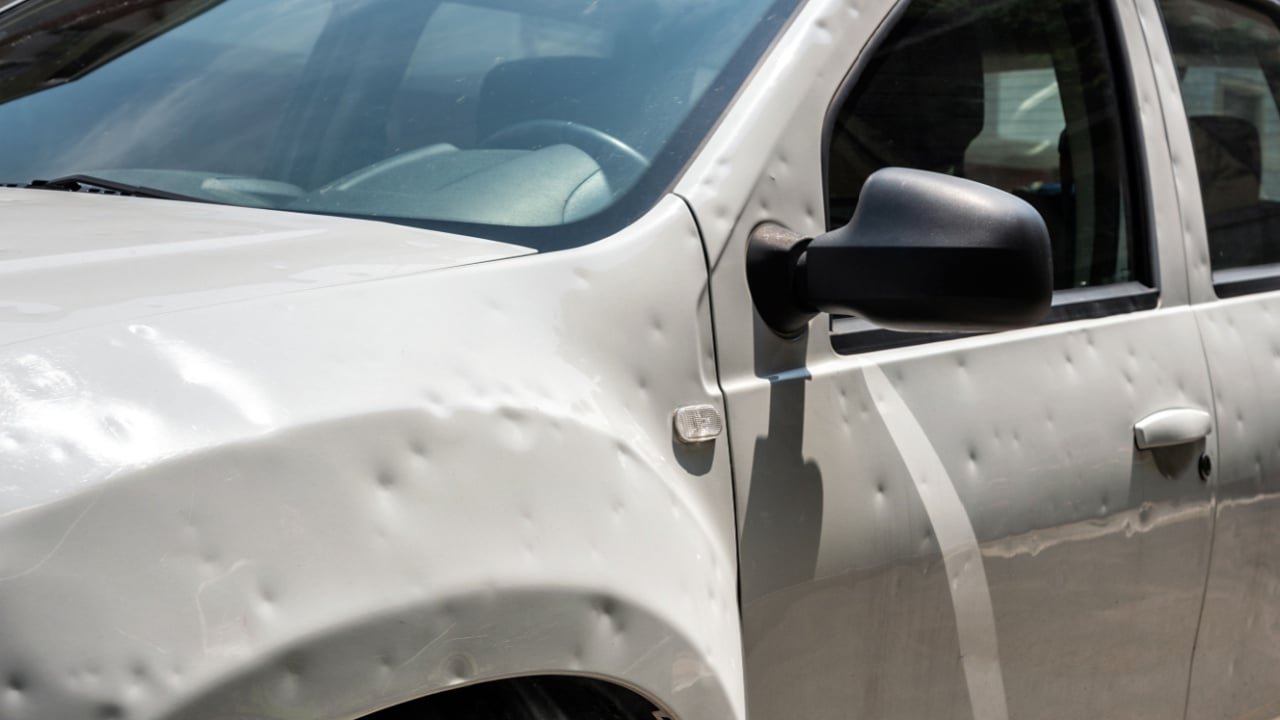 Damaged white car with many dents parked on city street on sunny day after hailstorm