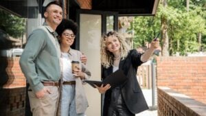 real estate agent pointing with pen near multiethnic couple standing near modern cottage