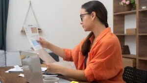 Concentrated woman takes out documents with utilities for payment from cardboard envelope sitting at table with laptop female studies papers in apartment
