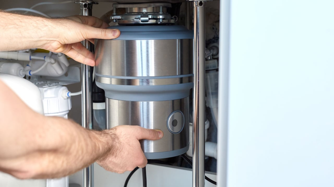 A plumber with gloved hands installs a household waste shredder for the kitchen sink. A technician installs or repairs a household waste shredder in the kitchen.