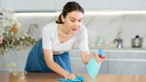 Young inexperienced housewife dressed in everyday clothes sprinkles cleaning agent on table in and polishes surface of countertop with piece of soft rag