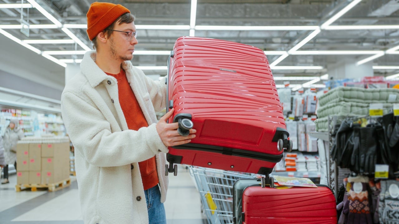Man in store chooses suitcase. Bag for travel, tourism, vacation. Young guy holding huge red suitcase, choosing, comparing. Huge supermarket, hypermarket with variety of goods for all occasions