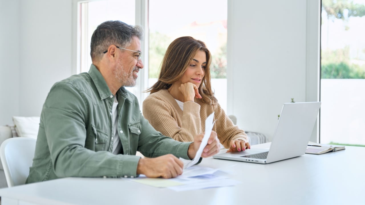 Happy middle aged mature man and woman paying bills online at home. Older senior couple using laptop computer checking insurance or financial invoice counting taxes sitting at table in living room.