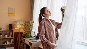Side view portrait of adult woman enjoying Spring cleaning at home and steaming curtains on window.
