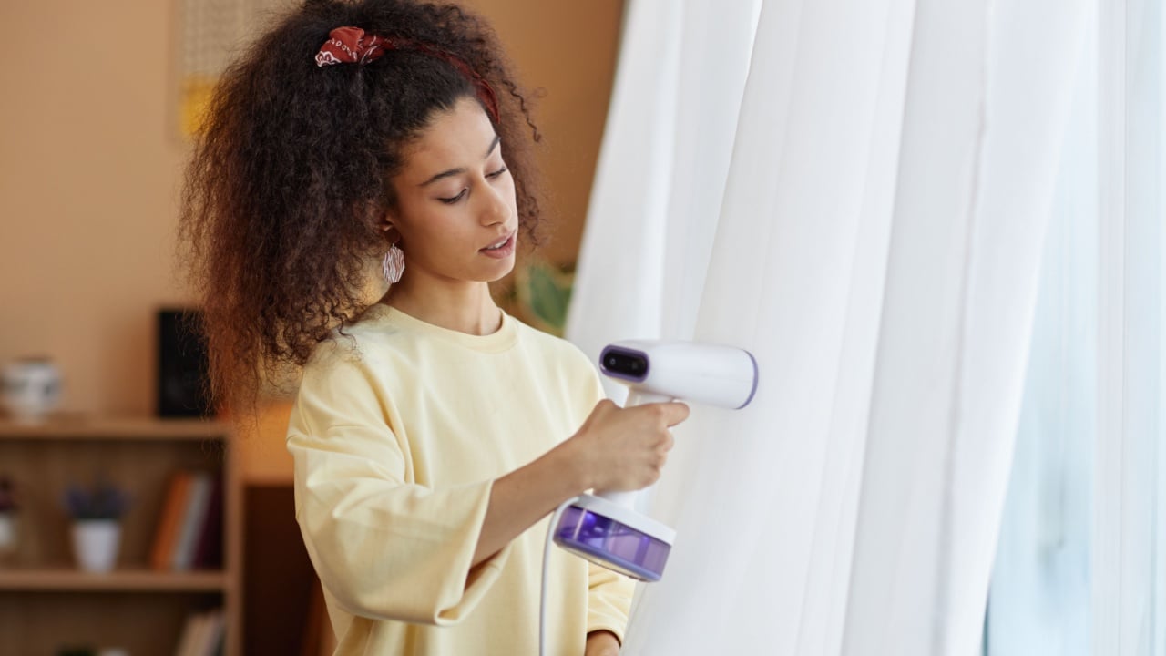 Waist up portrait of young woman steaming gauze curtains on window with with hand steamer during Spring cleaning copy space