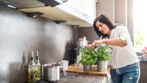 Beautiful mature woman taking care of herbs, cutting herbs for cooking in kitchen.