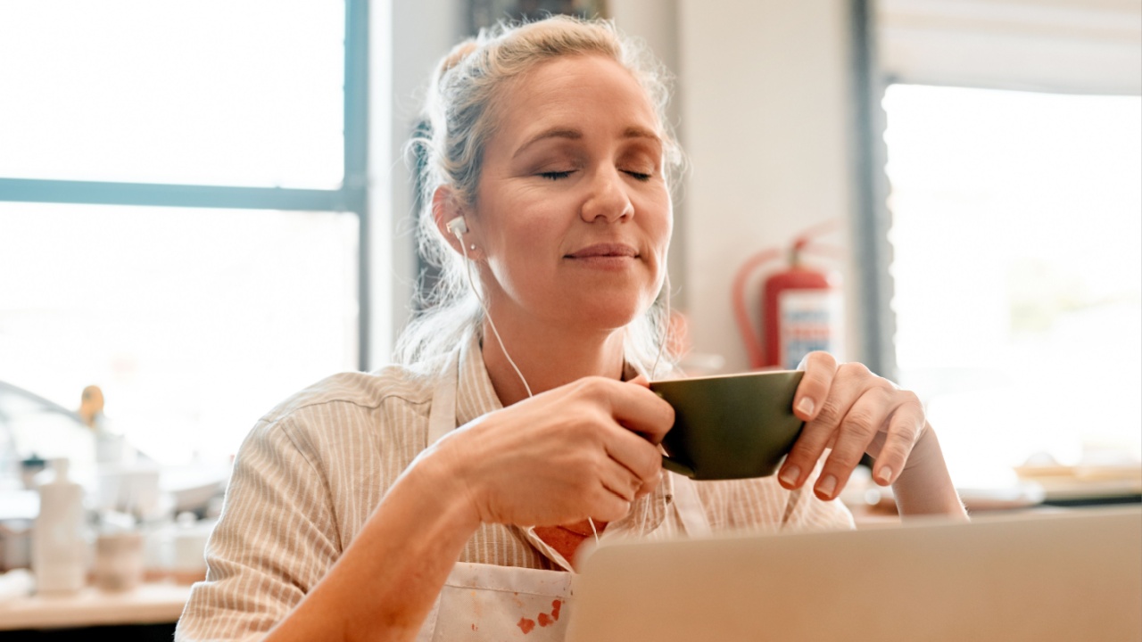Pottery, coffee and woman on laptop in workshop with earphones listening to artisan podcast for break. Calm, cappuccino and female potter with computer and audio tech for music in creative studio.