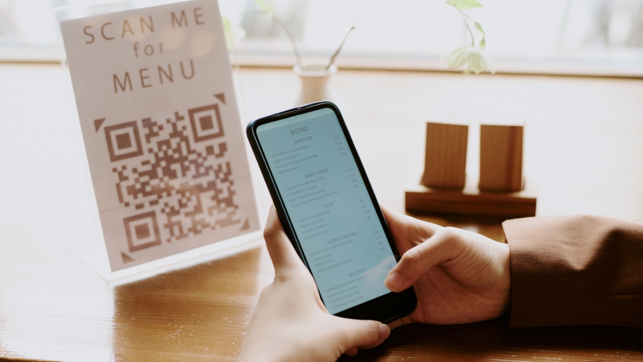 Person scanning menu QR code with smartphone in cozy restaurant setting, holding phone steadily while focusing on screen, ambient light reflecting on wooden table surface