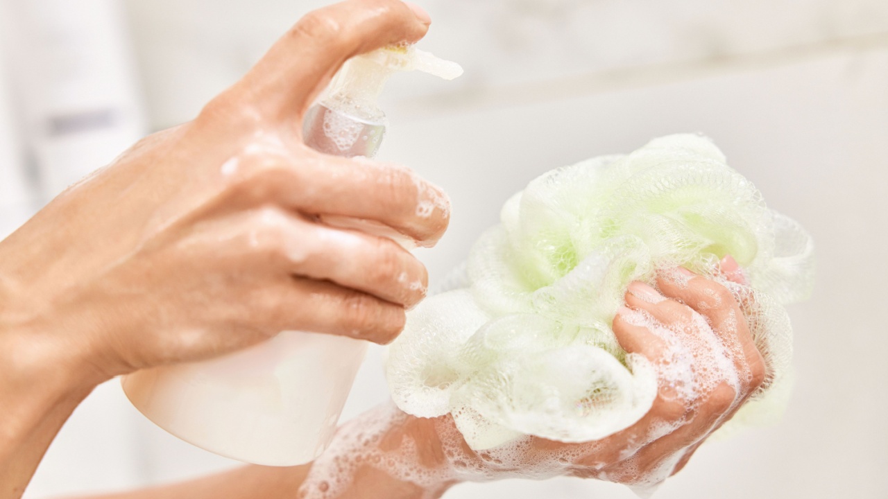 female hands applying liquid soap shower gel from white cosmetic bottle on sponge in bathroom