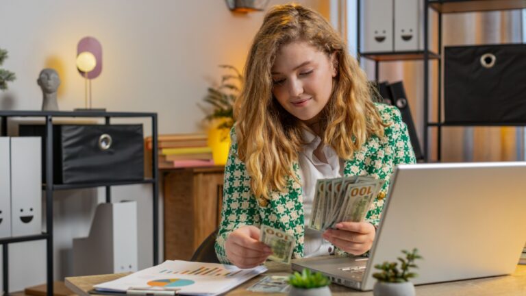 Happy Caucasian businesswoman counting money cash and using laptop in home office. Successful freelancer girl getting bundle of money in office, calculates dividends cash earnings profits and finances