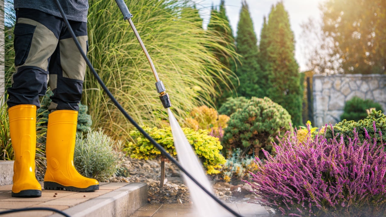 A person wearing yellow boots uses a pressure washer to clean a patio area surrounded by vibrant flowers and shrubs on a sunny afternoon.