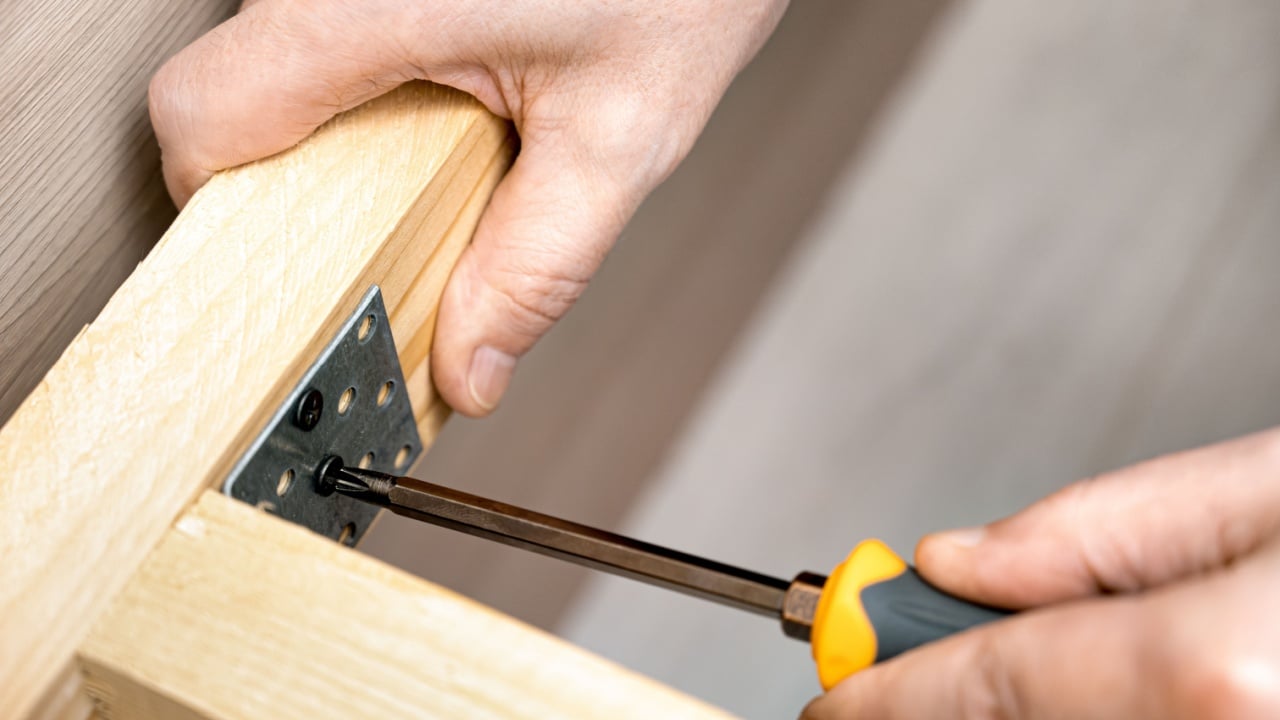Close-up of a carpenter's hands tightening a screw into a wooden frame with a screwdriver and metal bracket