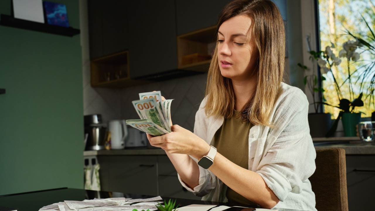 Focused woman counting dollars cash at kitchen table with receipts, notebook, and phone, organizing her personal finances. Concept of planning home budget