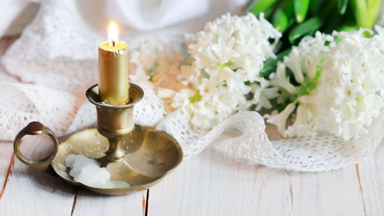 A brass candlestick with a gold candle next to a bouquet of white hyacinths on a wooden table.