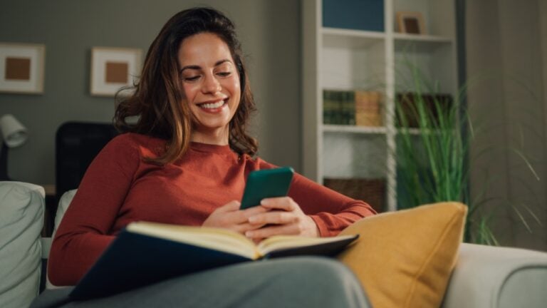 Happy woman relaxing on comfortable sofa holding mobile phone and reading book, enjoying leisure time in cozy living room, embracing technology and literature