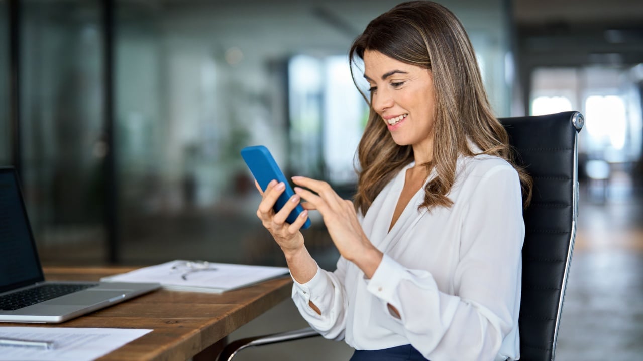 Happy business woman of middle age holding phone using cellphone in office. Smiling mature professional businesswoman executive using smartphone cell mobile apps on cellphone working sitting at desk.