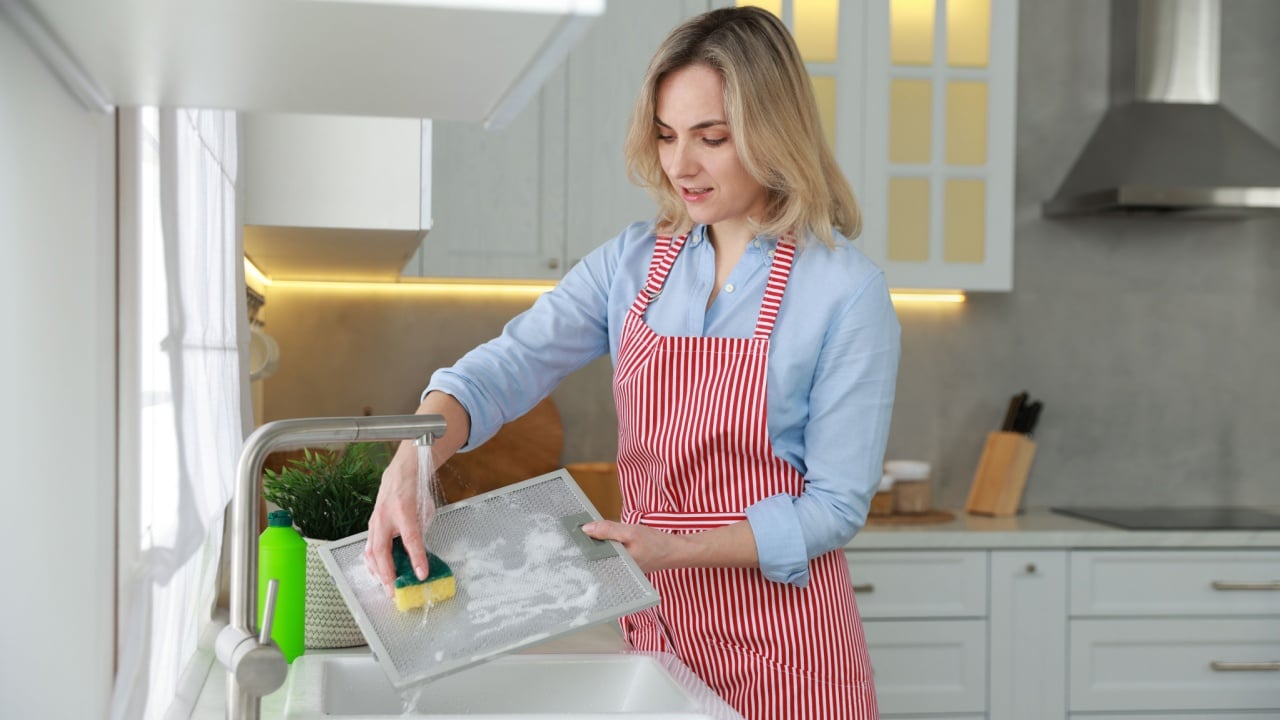 Woman cleaning filter of kitchen hood with sponge above sink indoors