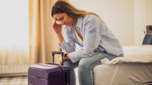 Woman in her 30s sitting on bed in hotel room. She is worried. Suitcase is beside the bed.