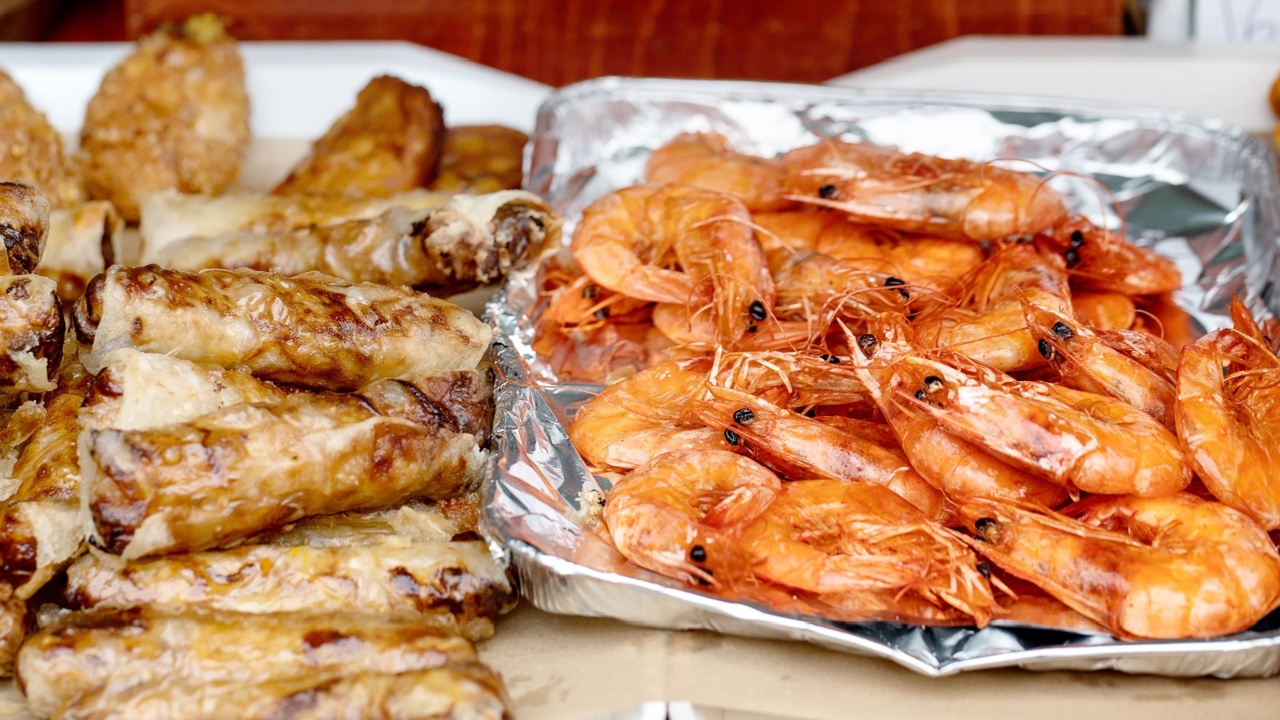 Delicious homemade spring rolls with meat filling served alongside cooked prawns in foil tray. Party food ready for serving on white plates against wooden background.