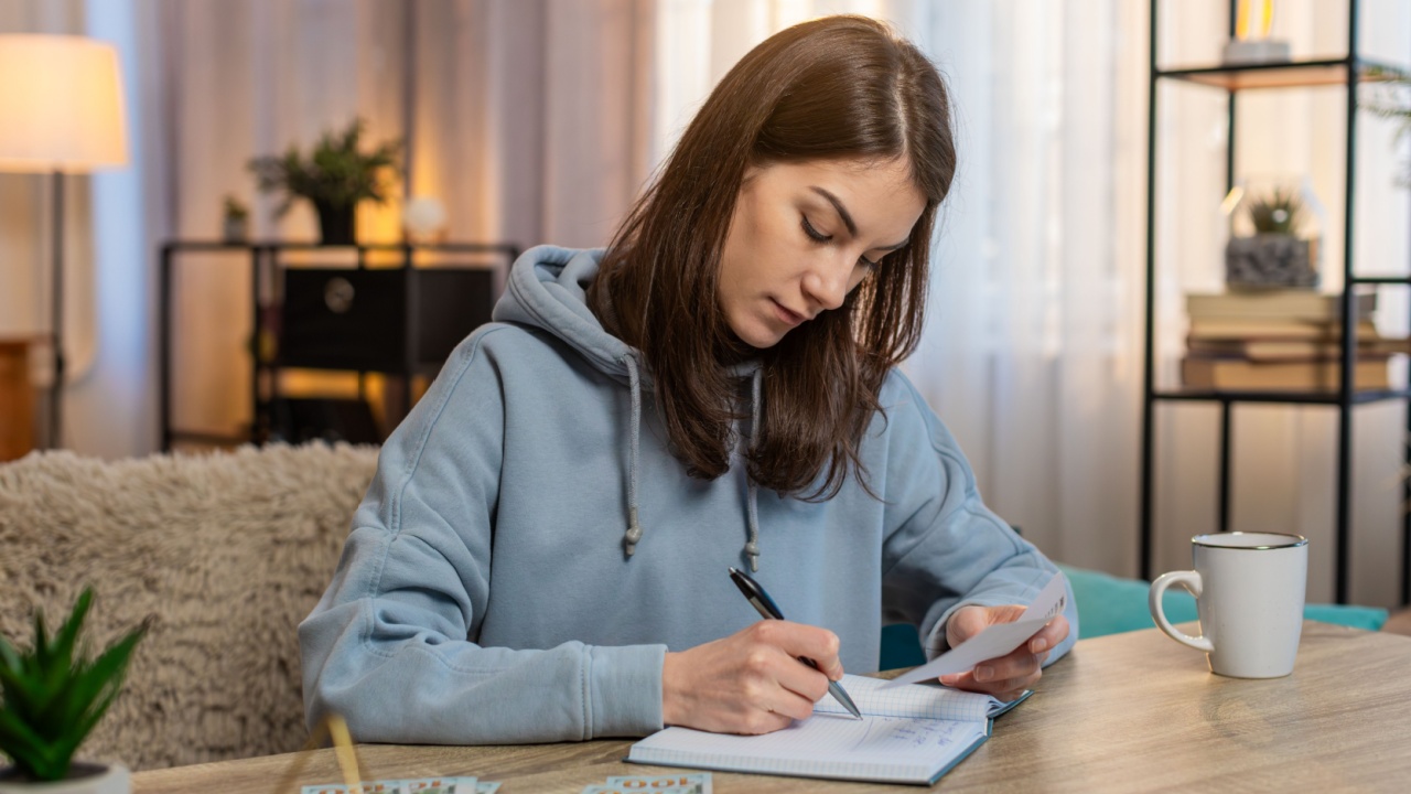 Focused young Caucasian woman making shopping list in notebook sitting at table on sofa looking at receipts and counting money in living room at home. Girl preparing notes for financial cost or budget