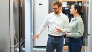 shop assistant demonstrates refrigerator to young woman in home appliance shopping mall supermarket.