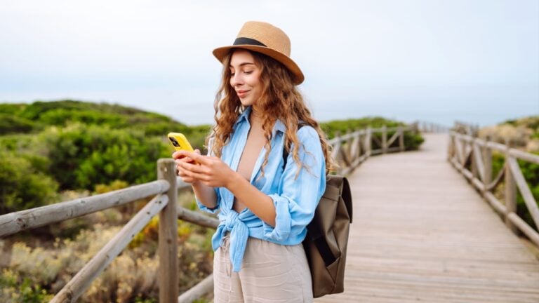 A cute tourist with a phone and a hat on a walking wooden path, blogging. Beautiful woman enjoying the seascape. Active lifestyle.