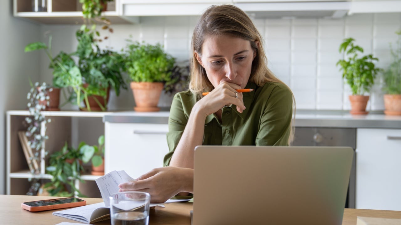 Pensive sorrowful woman reviewing bills with laptop and glass of water in home kitchen. Financial stress, debt issues, utility pay, household budget finance struggles, overdue accounts, money problems