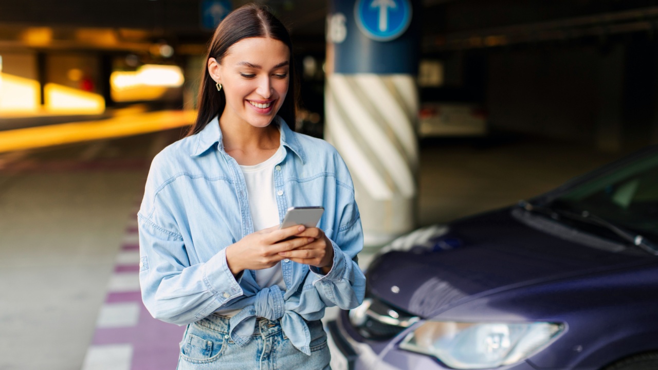 Happy lady using cellphone, standing near car on at underground parking, communicating or using online application. Car sharing, rental service or taxi app