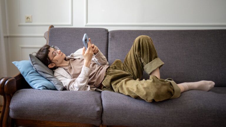 Procrastinating young woman lounging on sofa browsing chat apps on smartphone, laziness and emotional detachment. Apathy, digital dependency, indoor inertia, bad habit downtime, solitary existence