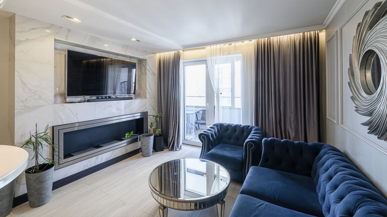 modern living room features a marble fireplace with a TV above, blue velvet seating, a mirrored coffee table, and curtains. Light flooring and a decorative mirror add to the room's elegance