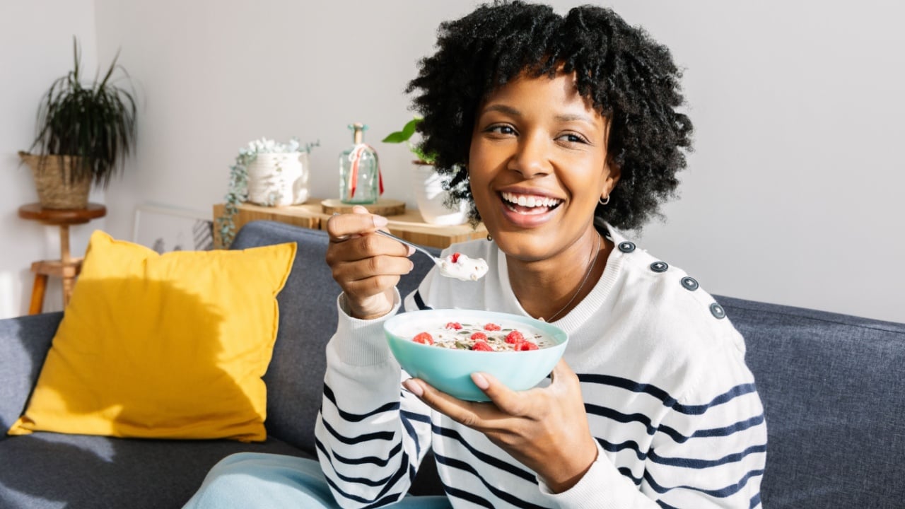Portrait of happy young Latin American woman enjoying healthy breakfast at home on the sofa on winter cozy day