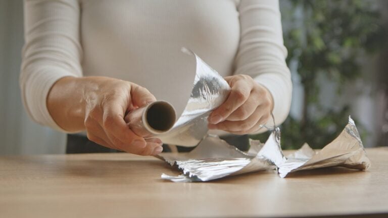 Close up of woman hands manipulating a shiny roll of aluminum foil on a wooden table, highlighting household activity.
